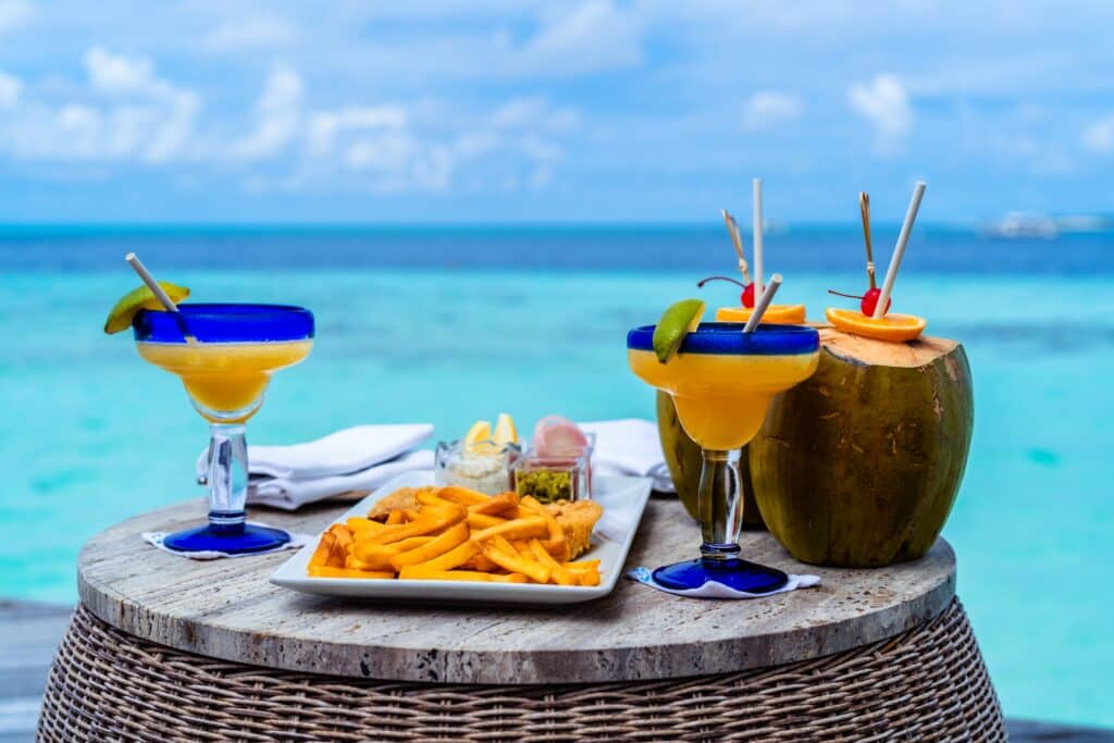Margaritas, some French fries, and some coconuts with straws in them in front of the beach.